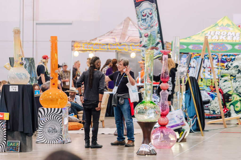 Attendees interact on the MJBizCon show floor in the Culture+ neighborhood, surrounded by large, colorful glass art and branded booths from exhibitors like Jerome Baker Designs.